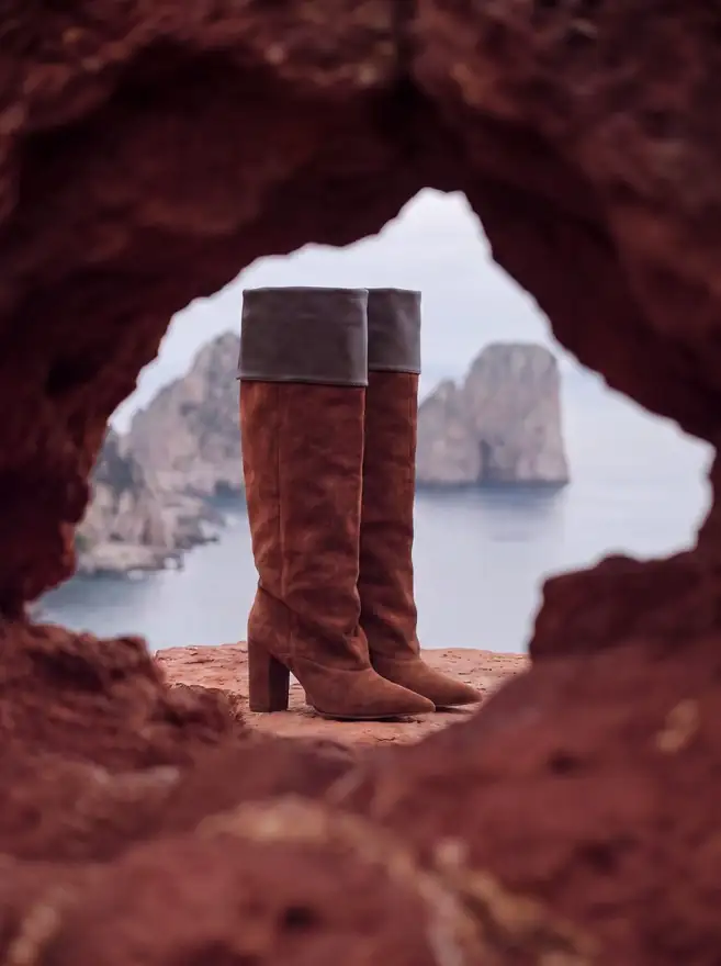Mascaró campaign image made with Ai: a pair of suede knee boots framed by a rock arch over the sea
